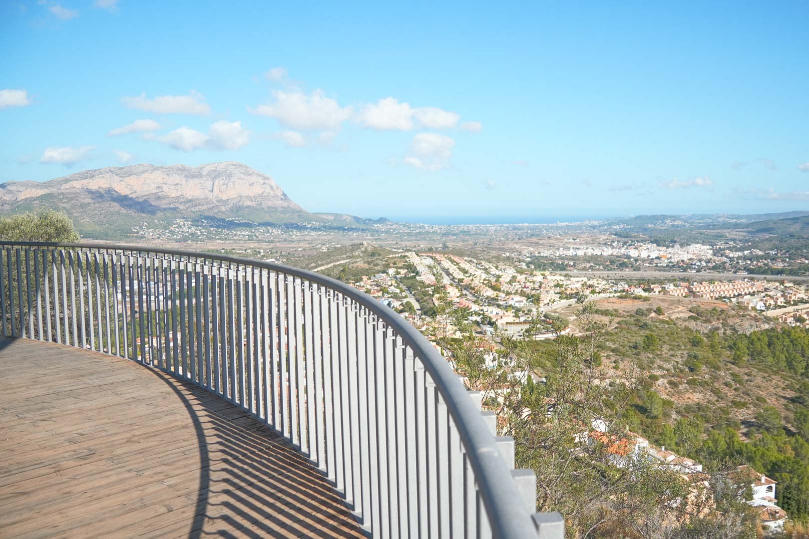 Geschwungene Holzterrasse mit Metallgeländer und Panoramablick über das Tal, die Berge und das Mittelmeer bei klarem Himmel.