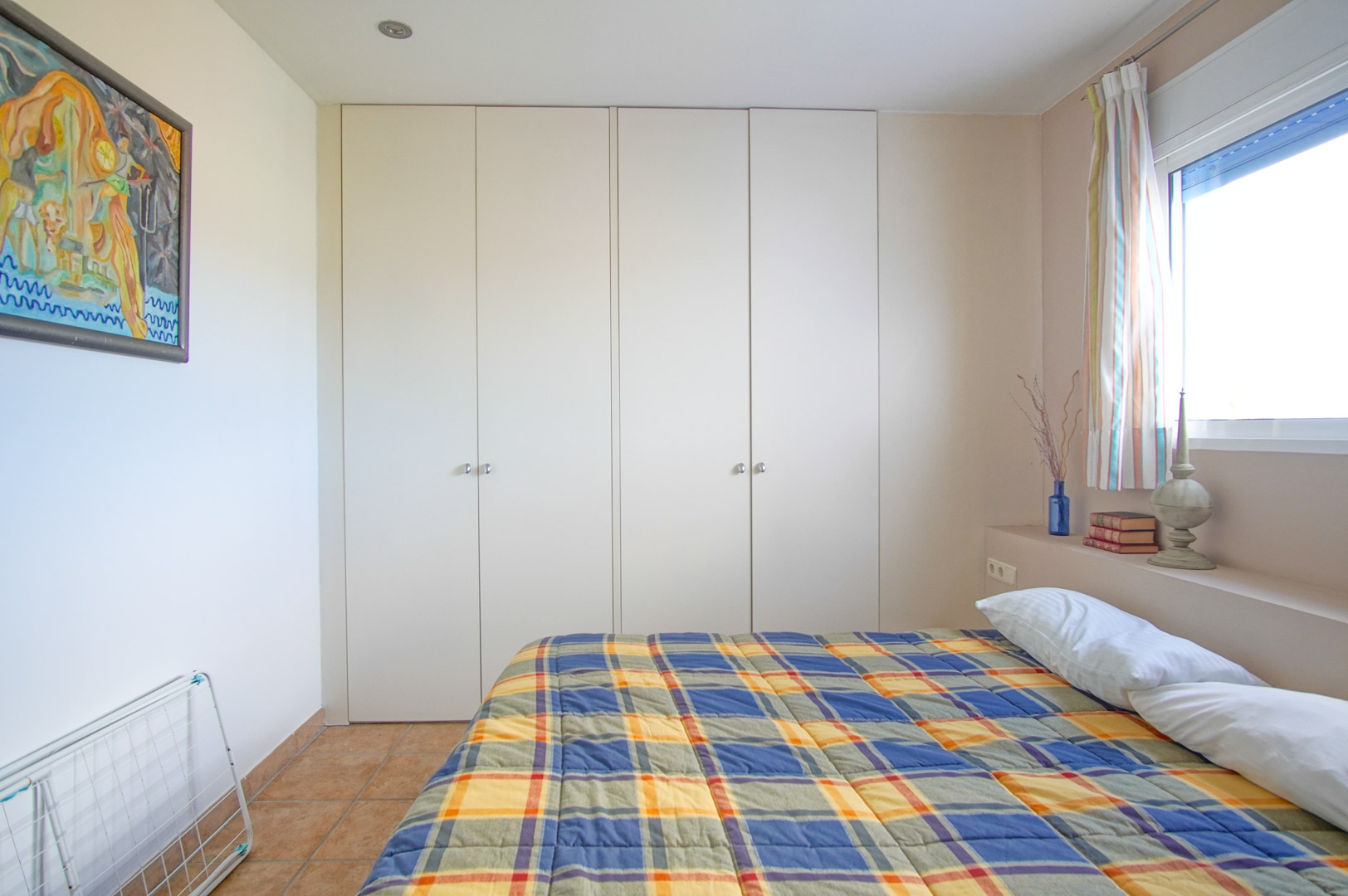 Helles Schlafzimmer mit großen weißen Einbauschränken, rustikalem Fliesenboden und einem Seitenfenster, das für reichlich natürliches Licht sorgt.