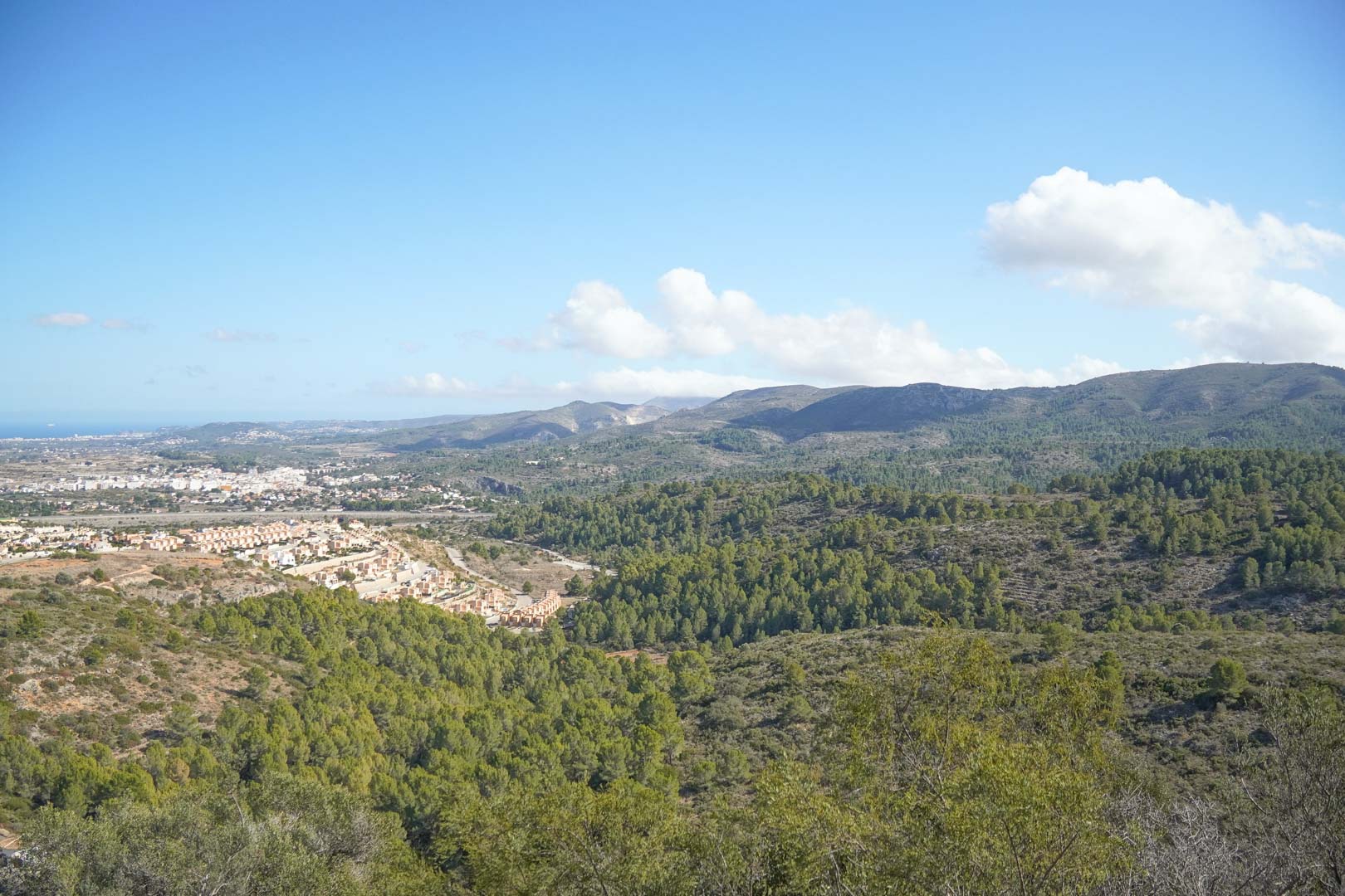 Panoramablick über das Pedreguer-Tal mit kiefernbewachsenen Bergen, Siedlungen und dem Mittelmeer am Horizont unter einem klaren, blauen Himmel.