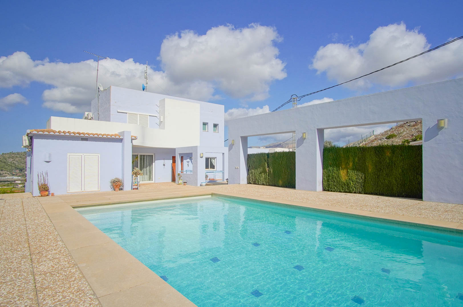 Rechteckiger Swimmingpool auf einer großzügigen Steinterrasse, flankiert von der modernen blau-weißen Fassade der Villa und architektonischen Wänden.