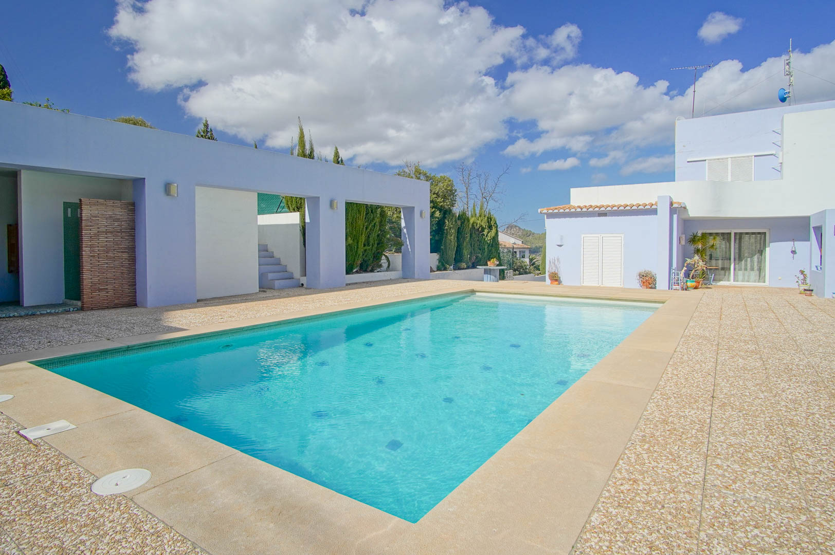 Rechteckiger Swimmingpool mit einer Terrasse aus Stein und Kies, umrahmt von lavendelfarbenen Außenwänden und mediterraner Bepflanzung.