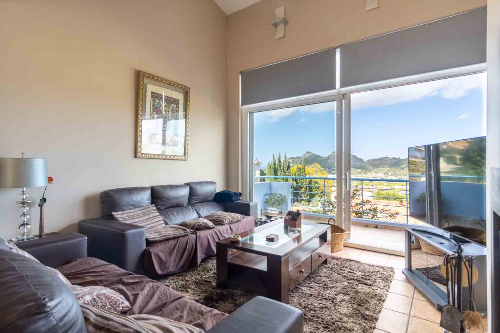 Wohnzimmer mit hohen Decken, dunklen Sofas und Holztisch. Große Glasschiebetüren führen auf den Balkon mit weitem Blick auf die Berge und das Tal.