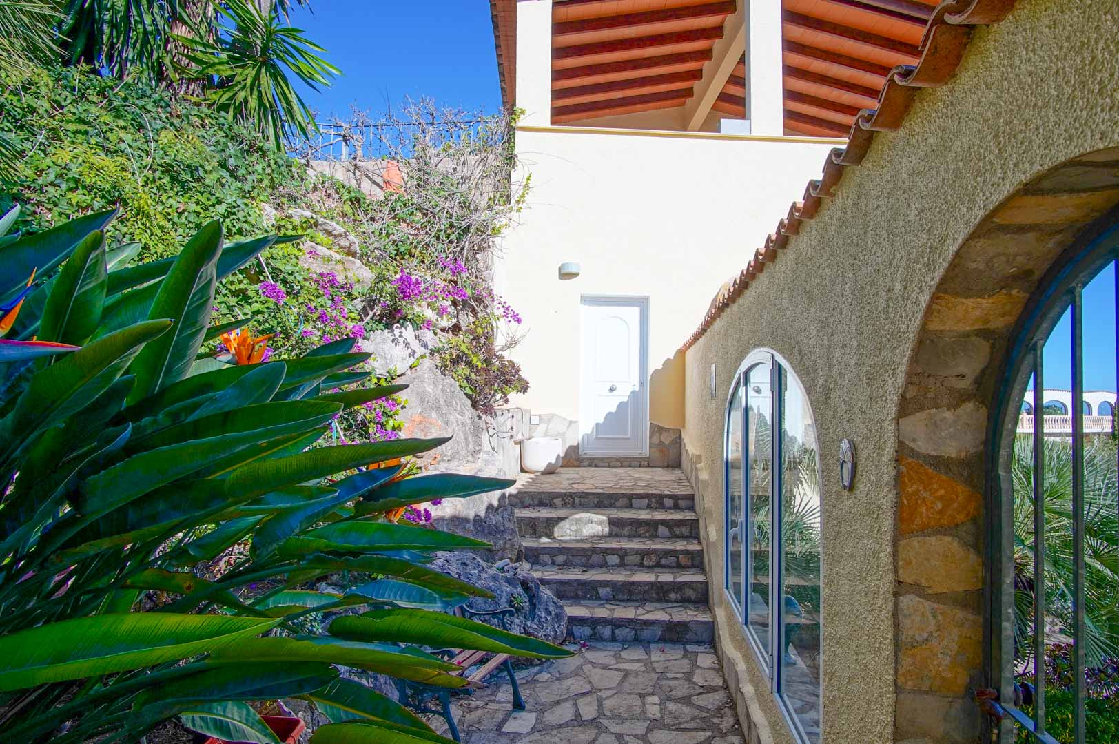 Außenbereich mit Natursteintreppen, mediterraner Bepflanzung und rustikalen Steinmauern mit Rundbogenfenstern.