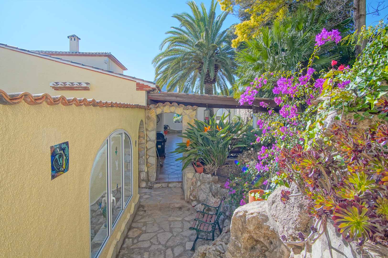 Außenweg aus Naturstein, gesäumt von Bougainvillea und Palmen, der durch einen rustikalen Steinbogen zu einem schattigen Terrassenbereich führt.