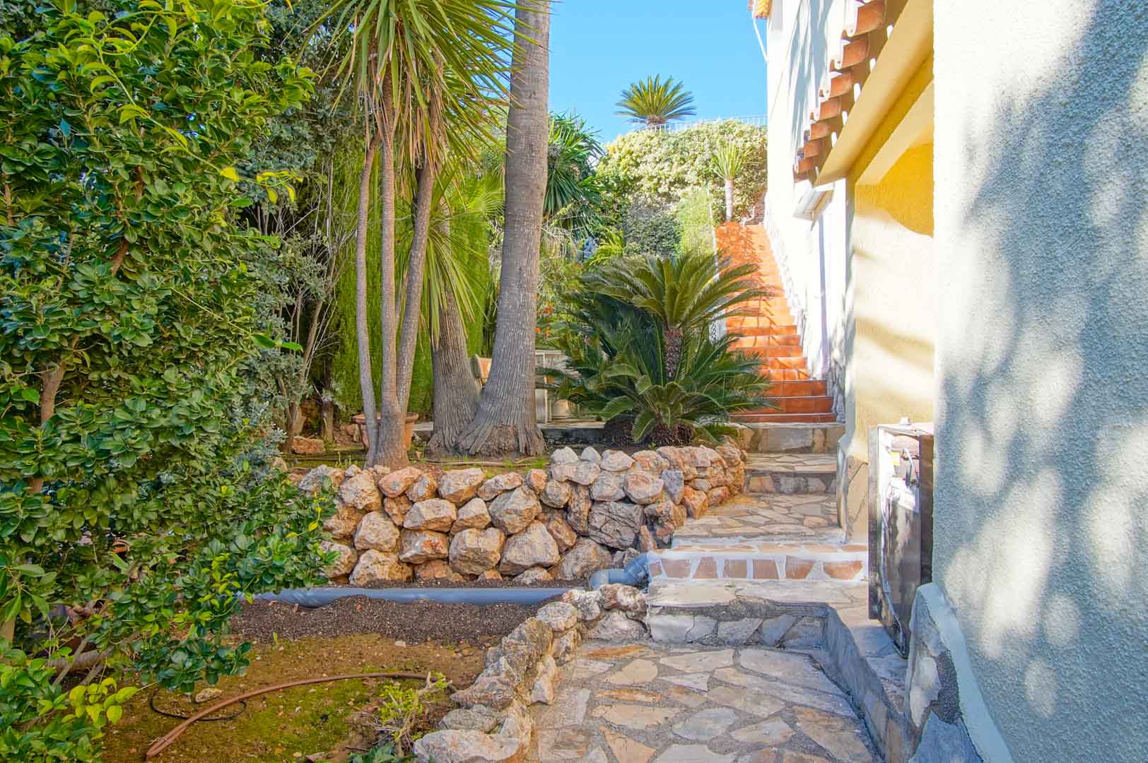 Außenweg aus Naturstein, gesäumt von Trockenmauern und mediterraner Bepflanzung, der zu einer Terrakotta-Treppe an der weißen Fassade der Villa führt.