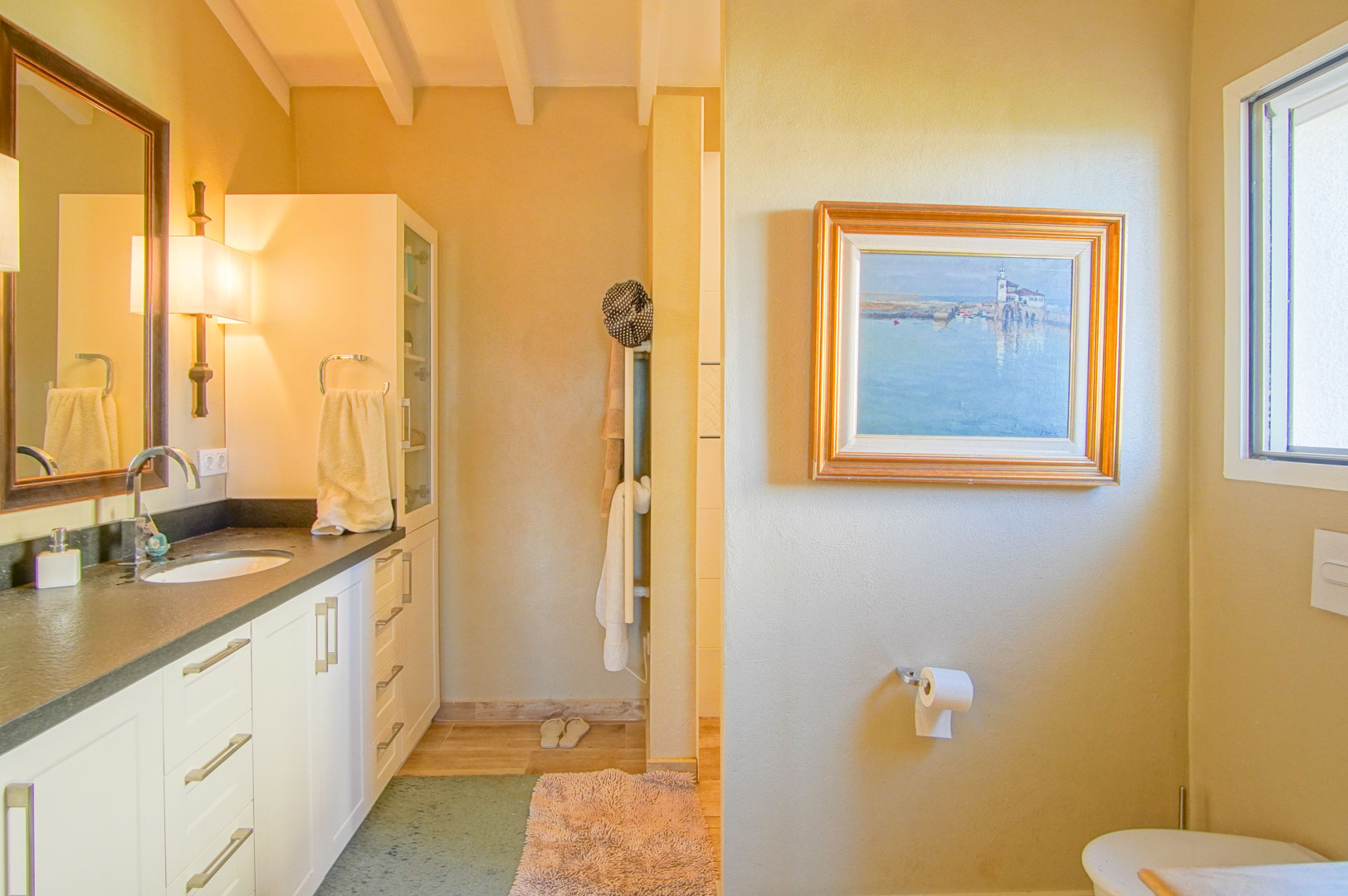 Badezimmer mit dunkler Arbeitsplatte, integriertem Waschbecken und sichtbaren Holzbalken. Es bietet weiße Schränke, warme Beleuchtung und natürliches Tageslicht.