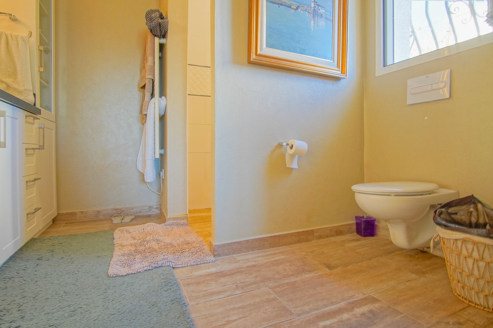 Badezimmer mit wandhängendem WC, Bodenfliesen in Holzoptik und weißen Schränken. Mit Handtuchheizkörper und Blick auf den gefliesten Duschbereich.