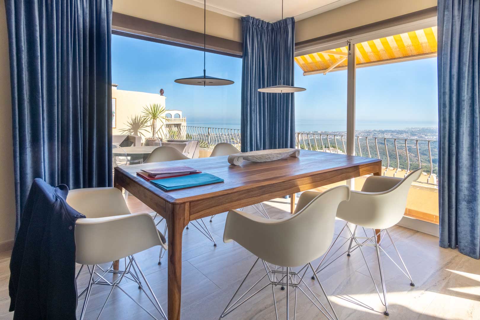 Essbereich mit Massivholztisch und Designerstühlen, umrahmt von bodentiefen Fenstern mit blauen Vorhängen und weitem Blick auf das Mittelmeer.