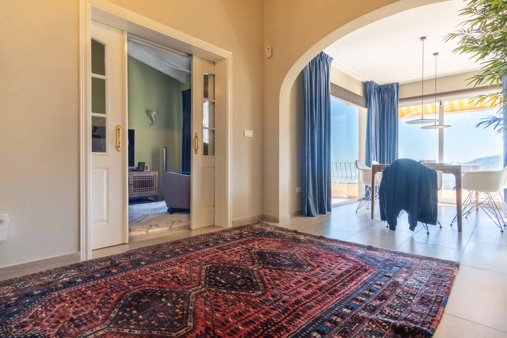 Flur mit Orientteppich, weißen Glasschiebetüren zum Wohnzimmer und einem Rundbogen, der in das Esszimmer mit weitem Meerblick führt.