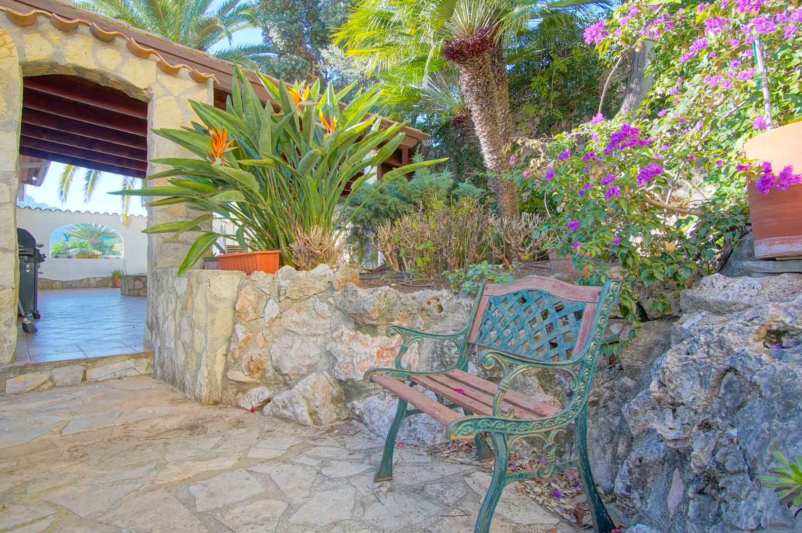 Gartenbereich mit klassischer Holzbank, Natursteinmauern und mediterraner Flora neben einer überdachten Terrasse mit markanten Holzbalken.