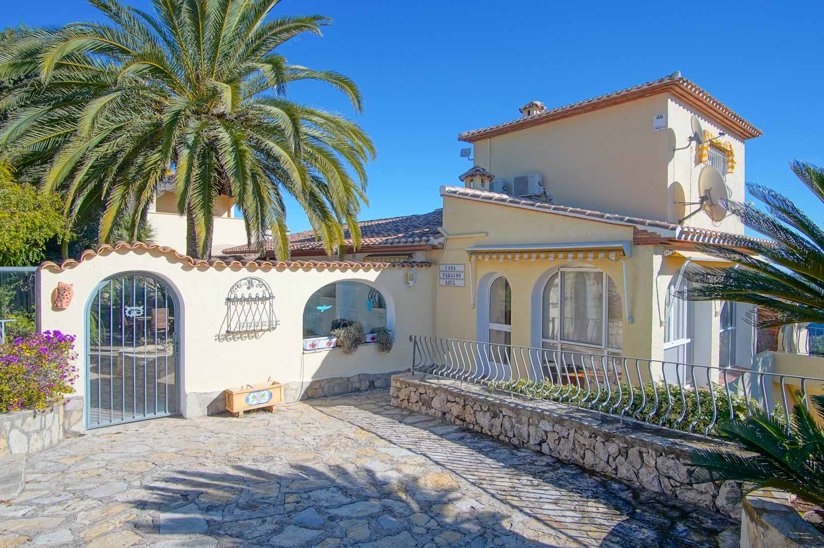 Hauptfassade der Villa mit gepflastertem Eingangsbereich, Natursteinmauern, dekorativen Rundbögen und mediterraner Bepflanzung bei klarem Himmel.