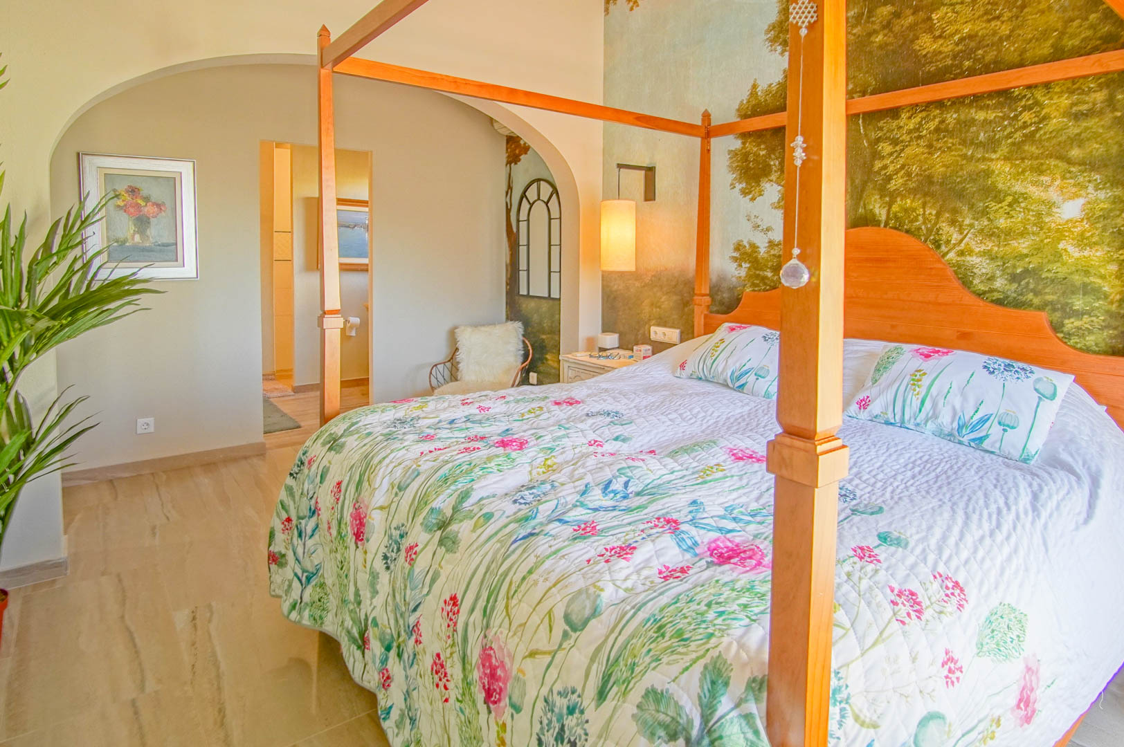 Hauptschlafzimmer mit Himmelbett aus Holz, Landschaftswandbild und poliertem Steinboden. Ein Rundbogen führt direkt zum angrenzenden Badezimmer.