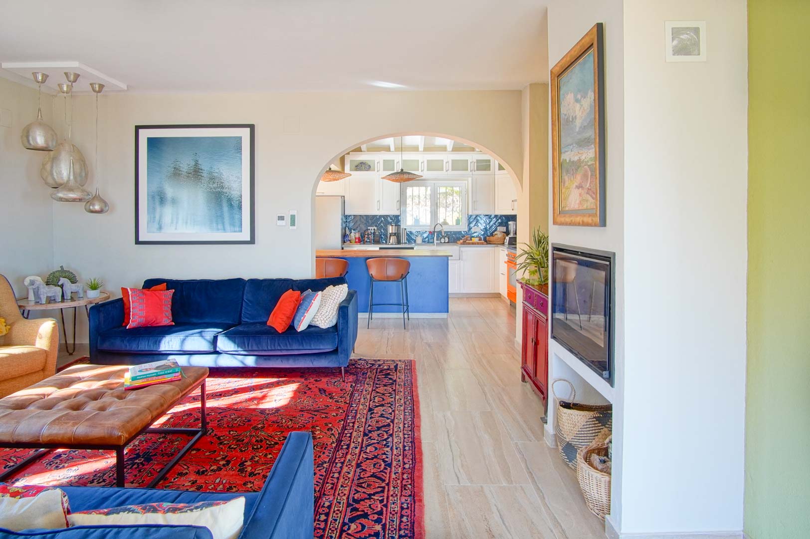 Heller Wohnbereich mit blauen Samtsofas und Einbaukamin, der durch einen Rundbogen in eine Küche mit blauer Kochinsel und weißen Schränken übergeht.