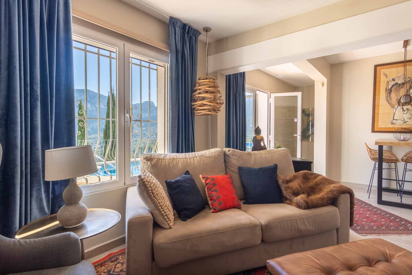 Heller Wohnbereich mit erdfarbenem Sofa, blauen Samtvorhängen und Fenstern mit Bergblick. Ein angrenzender Barbereich und ethnische Dekorelemente prägen den Raum.