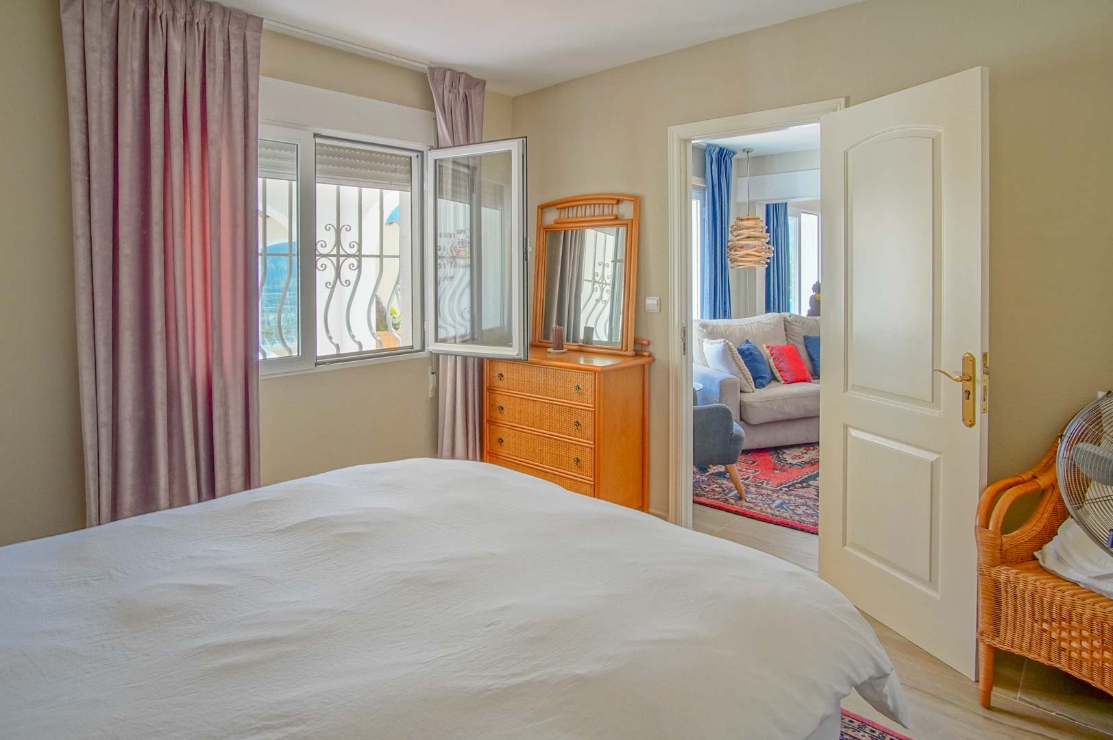 Helles Schlafzimmer mit Holzkommode und Fenster mit Ziergittern. Eine offene Tür führt in ein angrenzendes Wohnzimmer mit Sofa und gemustertem Teppich.