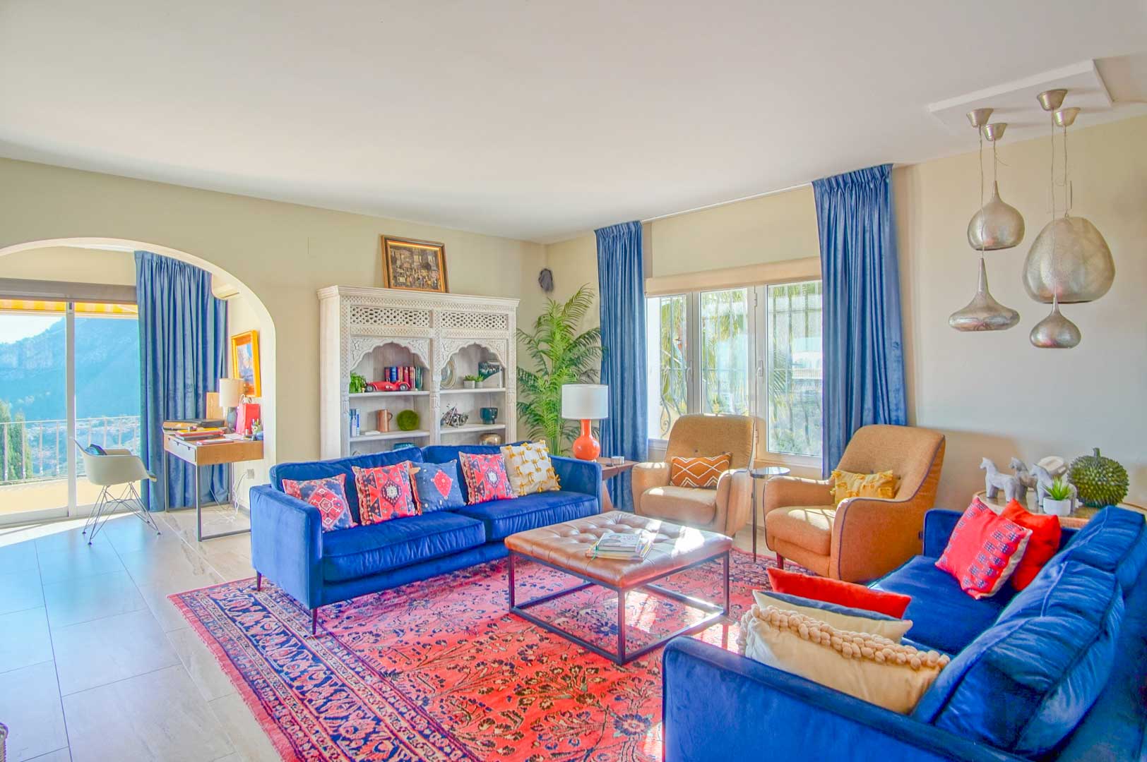 Helles Wohnzimmer mit blauen Samtsofas, Orientteppich und geschnitztem weißem Regal. Ein Rundbogen führt zum Arbeitsbereich mit Bergblick.