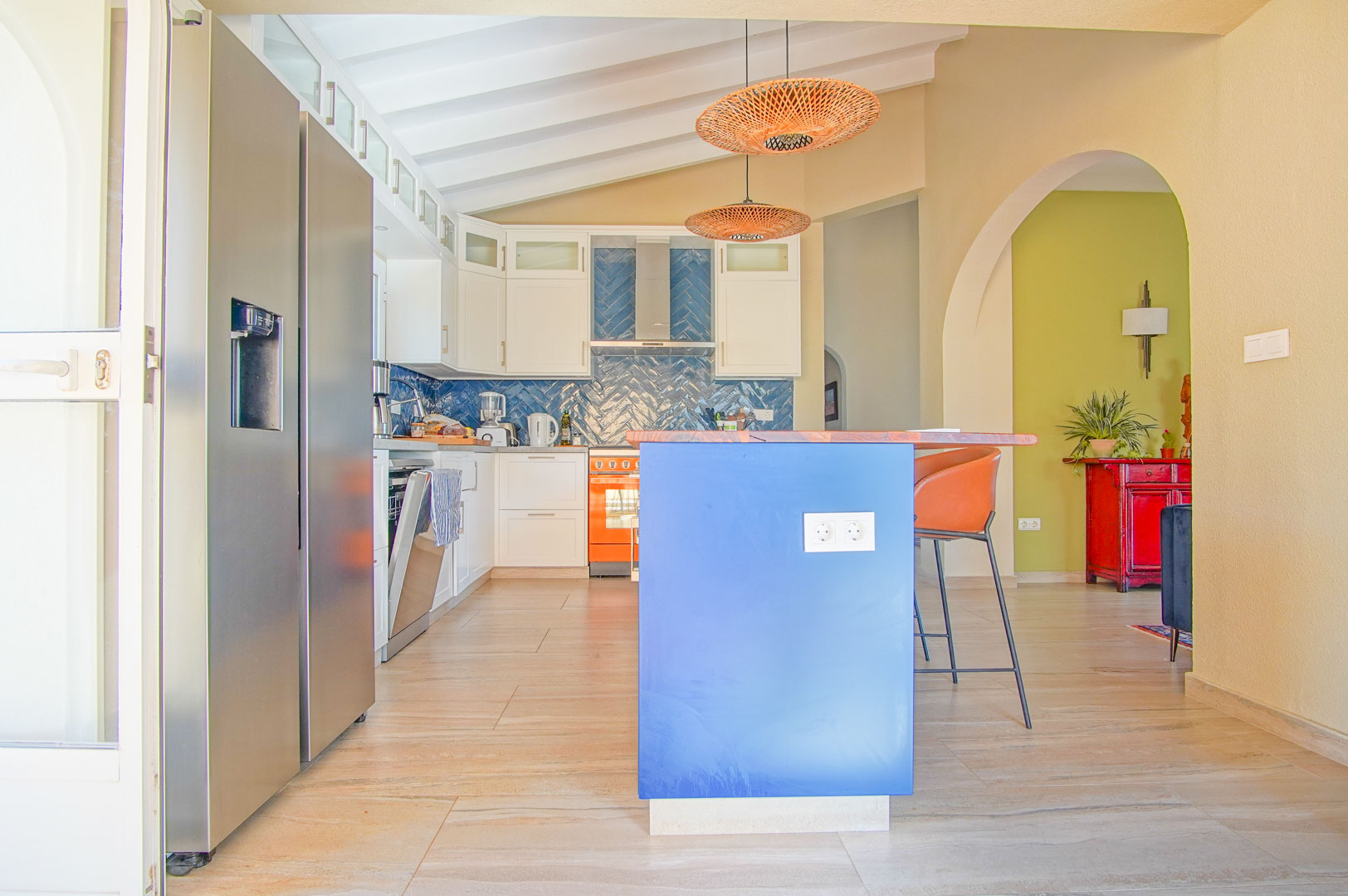Moderne Küche mit blauer Insel, Holzplatte und Fischgrät-Fliesenspiegel. Weiße Balkendecke, Rattanhängeleuchten und ein Side-by-Side-Kühlschrank prägen den Raum.