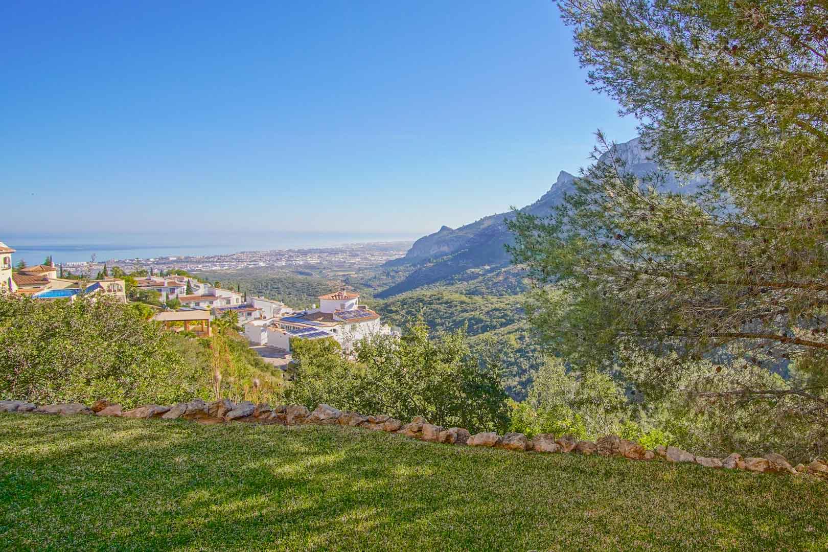 Panoramablick vom gepflegten Rasen auf die Mittelmeerküste und die Gebirgskette, eingerahmt von Pinien und mediterraner Vegetation.