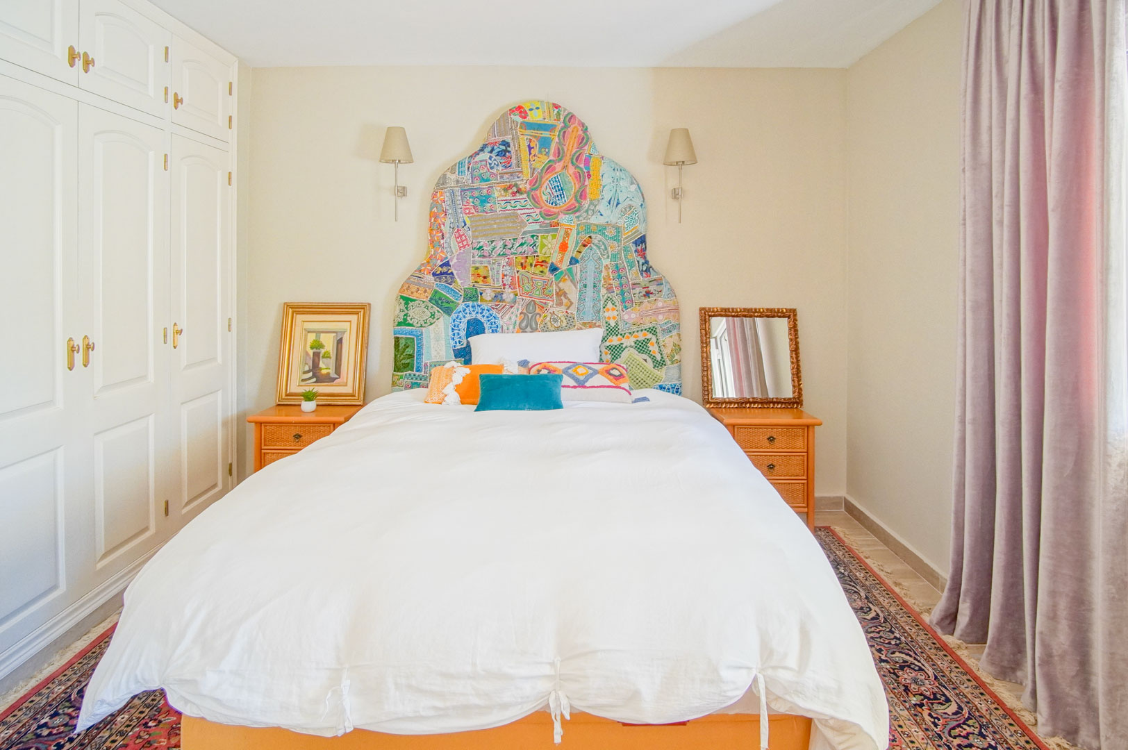 Schlafzimmer mit farbenfrohem Patchwork-Kopfteil, weißen Einbauschränken und Nachttischen aus Holz. Wandleuchten und ein Orientteppich setzen klassische Akzente.