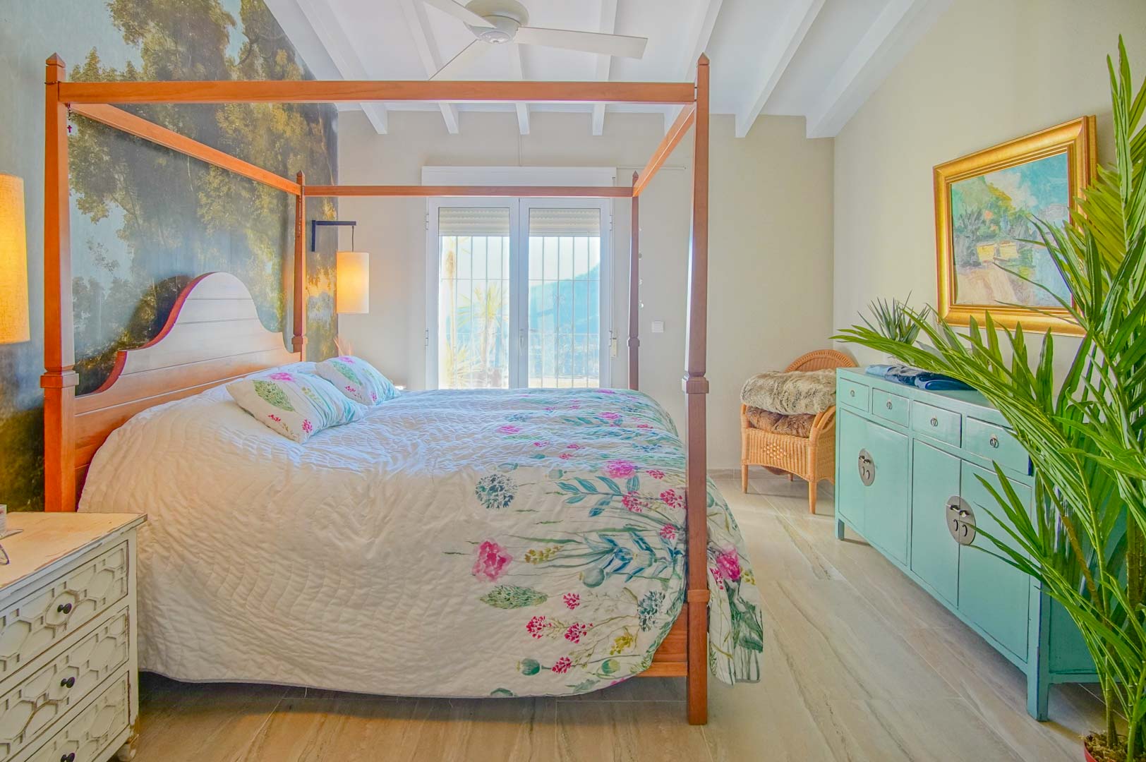 Schlafzimmer mit hölzernem Himmelbett, Wald-Wandbild, weißen Deckenbalken und Zugang zum Balkon durch verglaste Türen.
