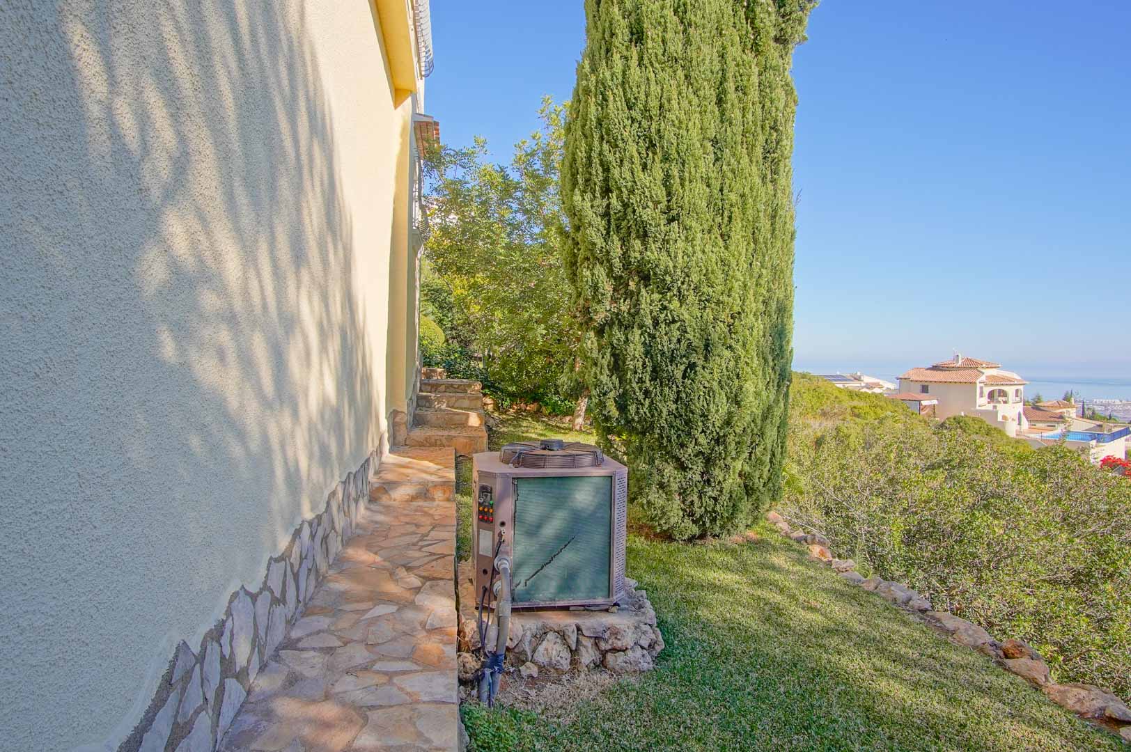 Seitlicher Natursteinweg entlang der strukturierten Fassade mit weitem Meerblick und mediterraner Bepflanzung inklusive einer hohen Zypresse.
