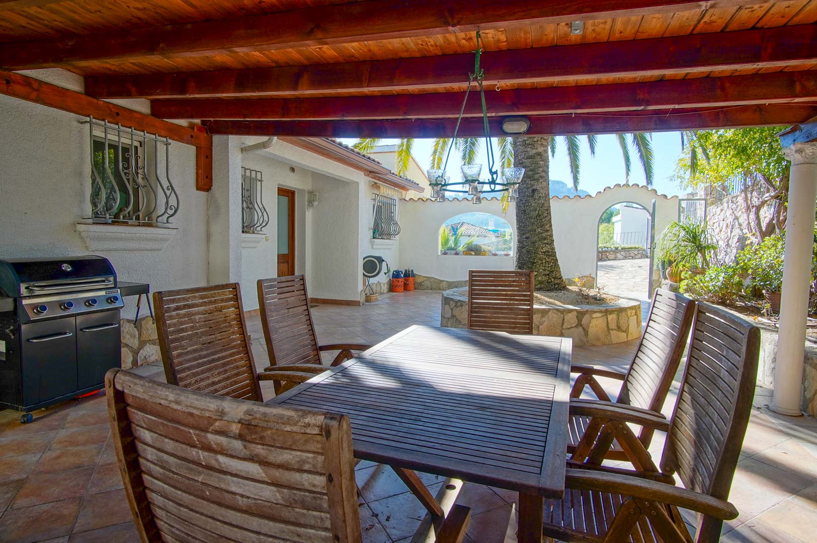 Überdachte Terrasse mit Holzbalkendecke, Essbereich im Freien, Gasgrill und ein Innenhof mit Palme und Natursteinmauern.