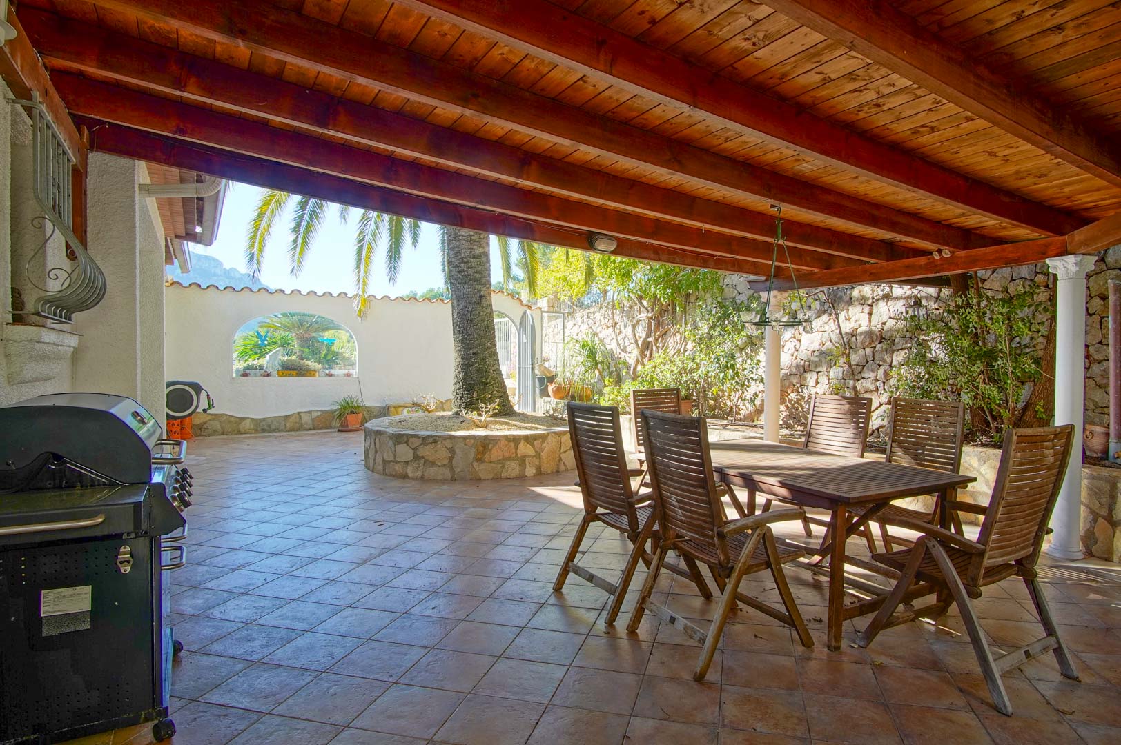 Überdachte Terrasse mit Holzbalkendecke, Essbereich im Freien, Grillstation und kreisförmigem Steinbeet mit Palme vor Natursteinmauern.
