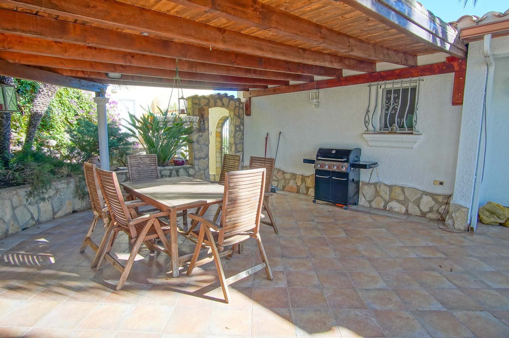 Überdachte Terrasse mit Holzbalkendecke, Essbereich im Freien und Gasgrill, ausgestattet mit Fliesenboden und Natursteinsockel.