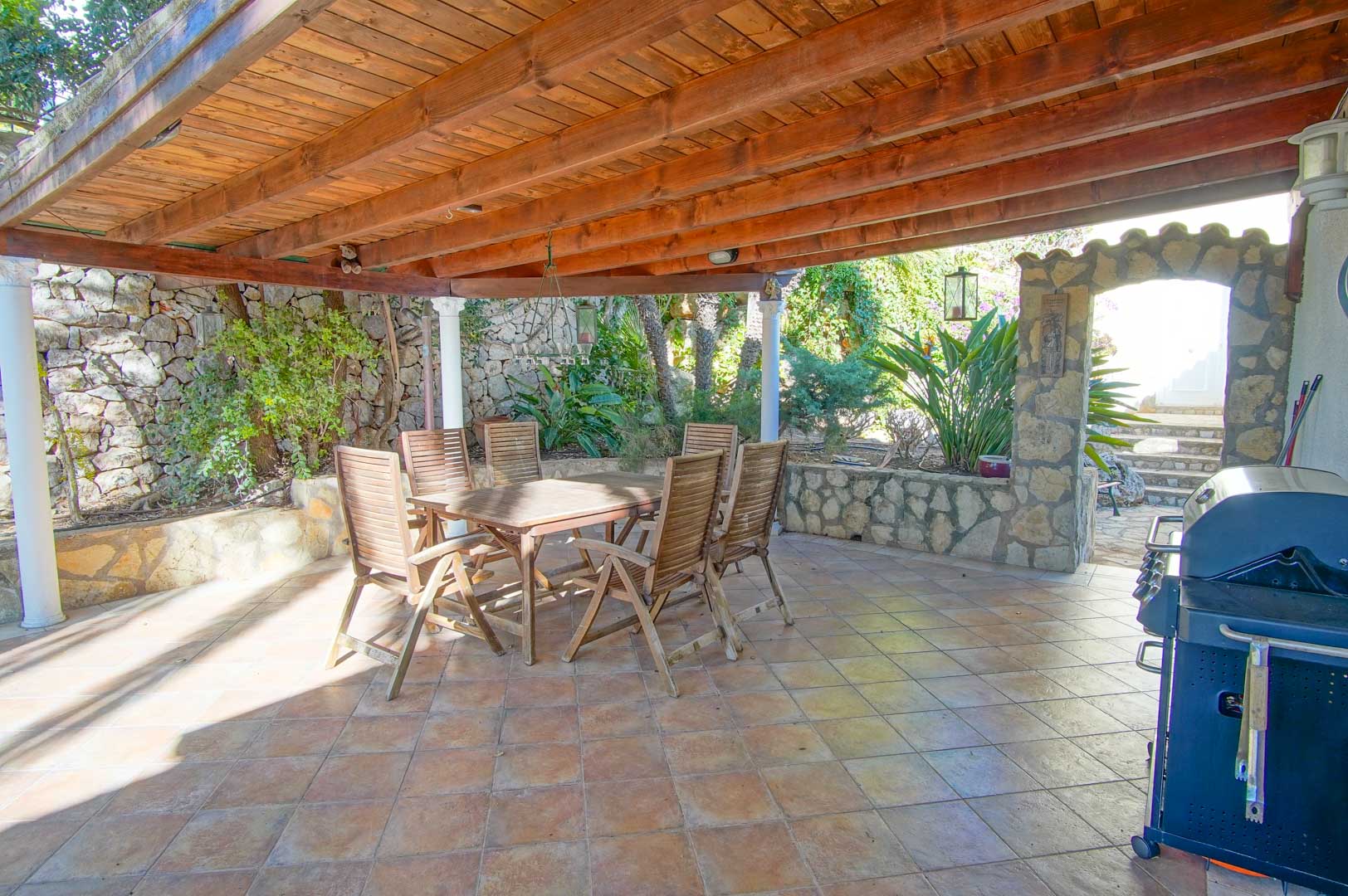 Überdachter Außenessbereich mit einer massiven Holzpergola, Natursteinmauern, Grillstation und einer rustikalen Sitzgruppe aus Holz.
