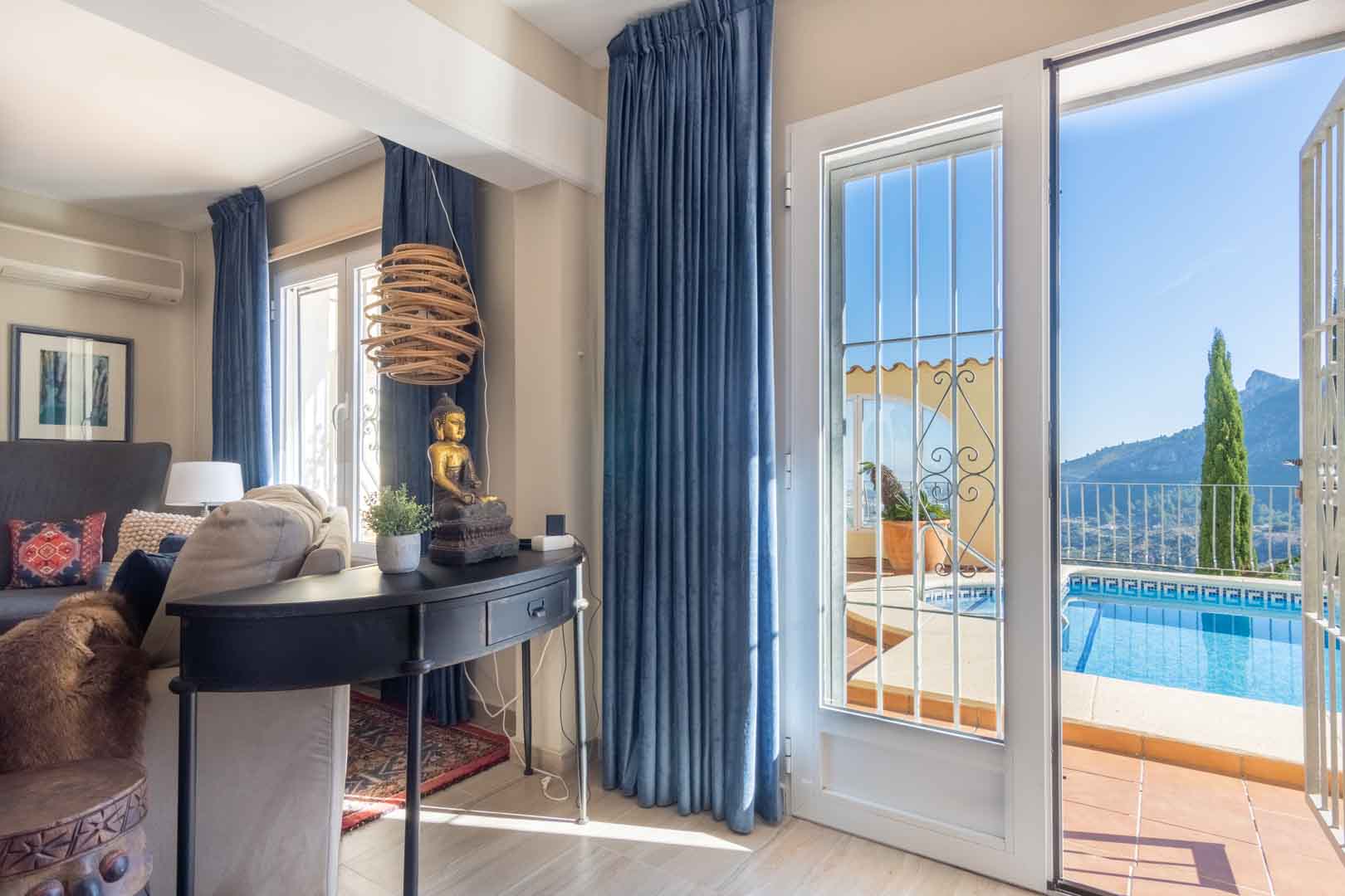 Wohnbereich mit blauen Samtvorhängen und Zugang zur Poolterrasse, die einen weiten Blick auf die Berge und die mediterrane Umgebung bietet.