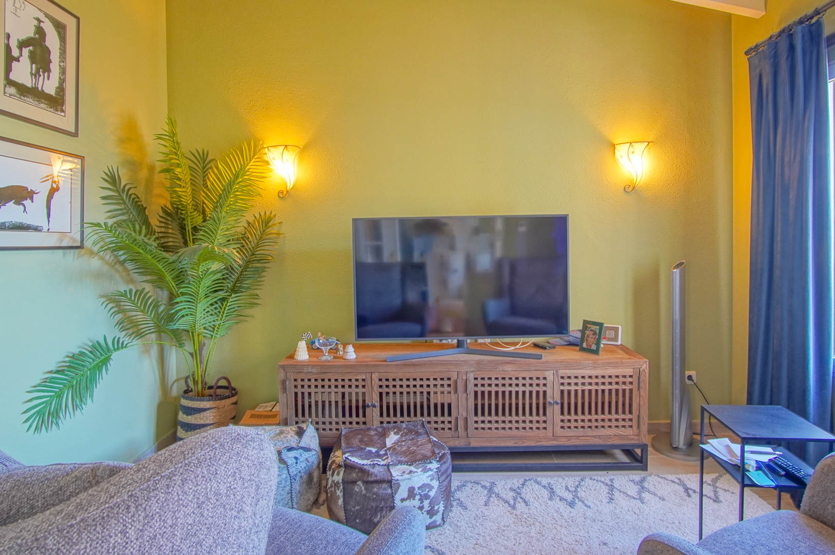 Wohnbereich mit TV-Kommode aus Holz im Gitterdesign, warmen Wandleuchten, Zimmerpflanze und gemütlichen Sitzhockern auf einem gemusterten Teppich.