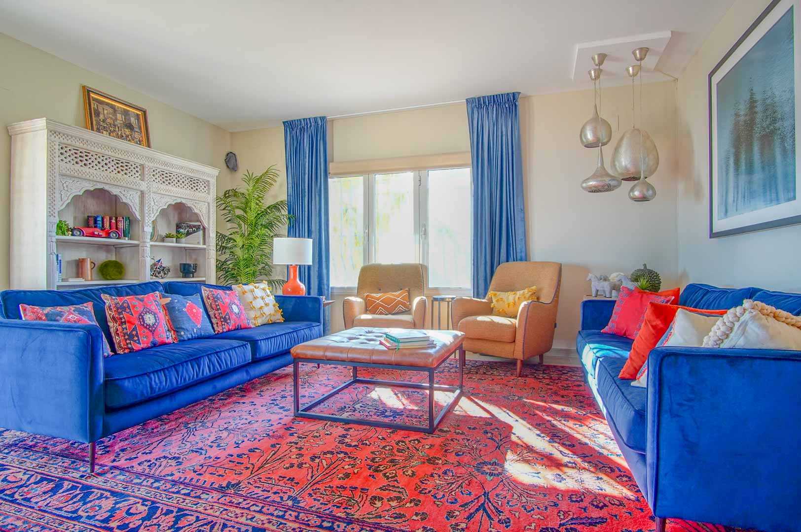 Wohnzimmer mit blauen Samtsofas, rotem Orientteppich und einem weißen geschnitzten Regal. Natürliches Licht und silberne Pendelleuchten prägen den Raum.