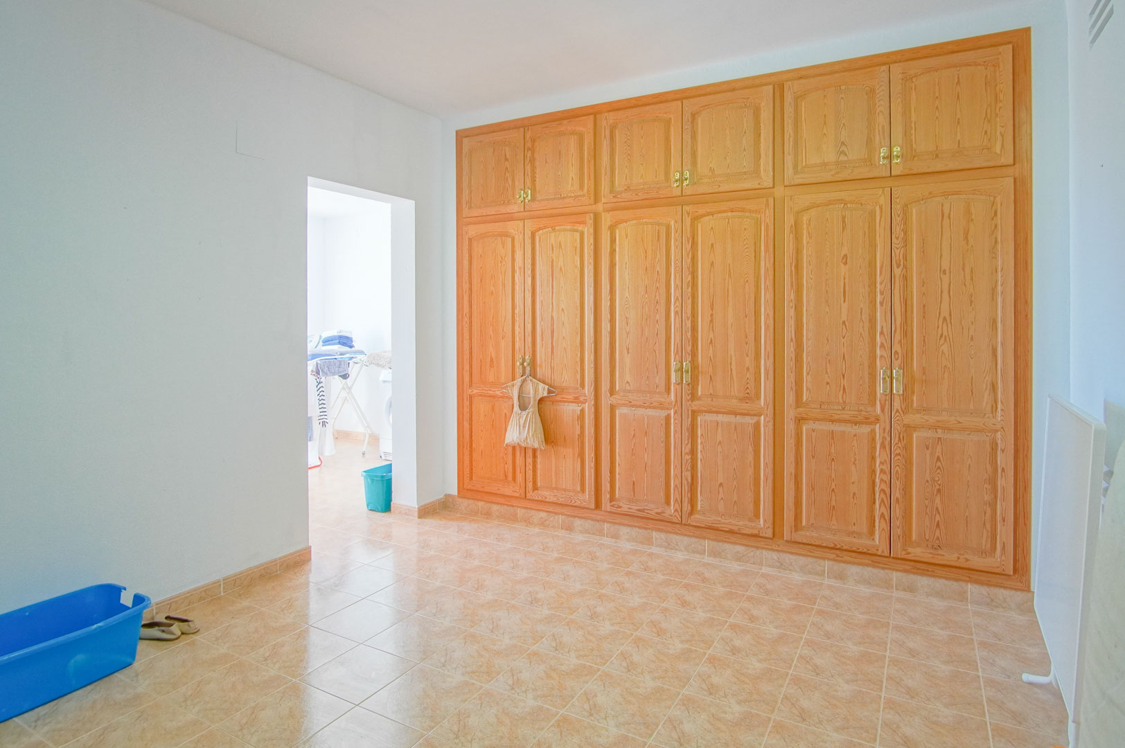 Zimmer mit großem Einbauschrank aus Holz, Fliesenboden und Durchgang zum angrenzenden Waschraum mit natürlicher Belichtung.