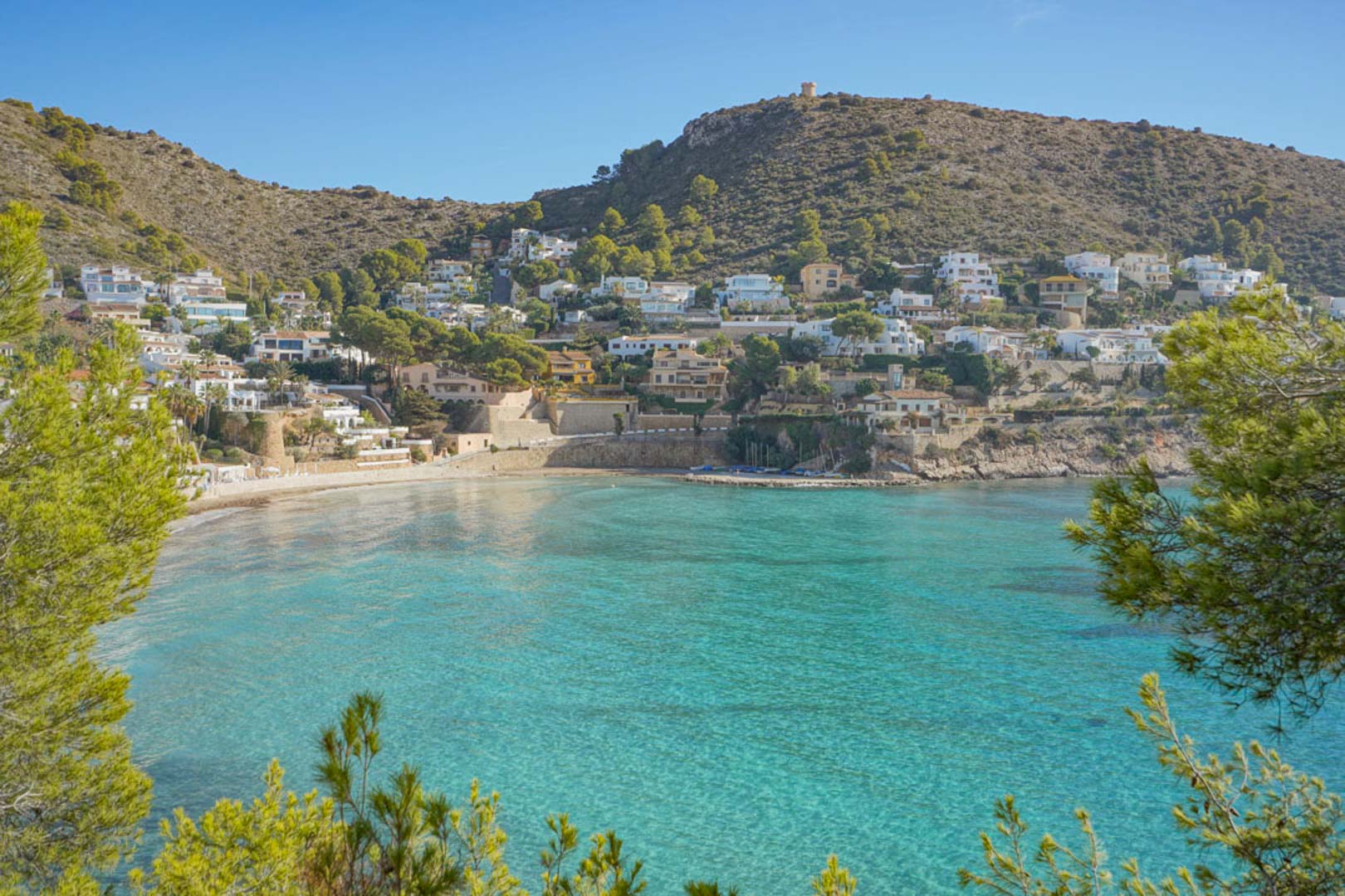 Panoramablick auf die Bucht von El Portet in Moraira mit kristallklarem, türkisfarbenem Wasser, umgeben von Pinien und exklusiven Villen am Hang.