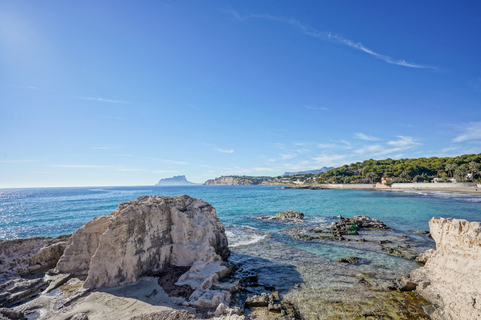 Panoramablick auf die Küste von Moraira mit natürlichen Felsformationen, kristallklarem türkisfarbenem Wasser und dem am Horizont sichtbaren Peñón de Ifach.