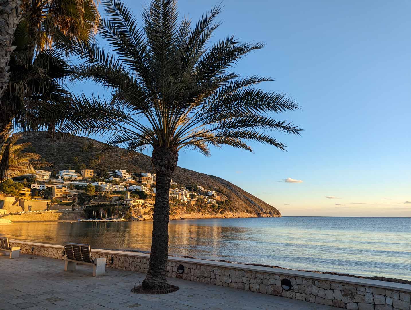Steinpflaster-Küstenpromenade in Moraira mit Palmen und Holzbänken, mit Blick auf das ruhige Mittelmeer und die villenbesetzten Hänge im Abendlicht.