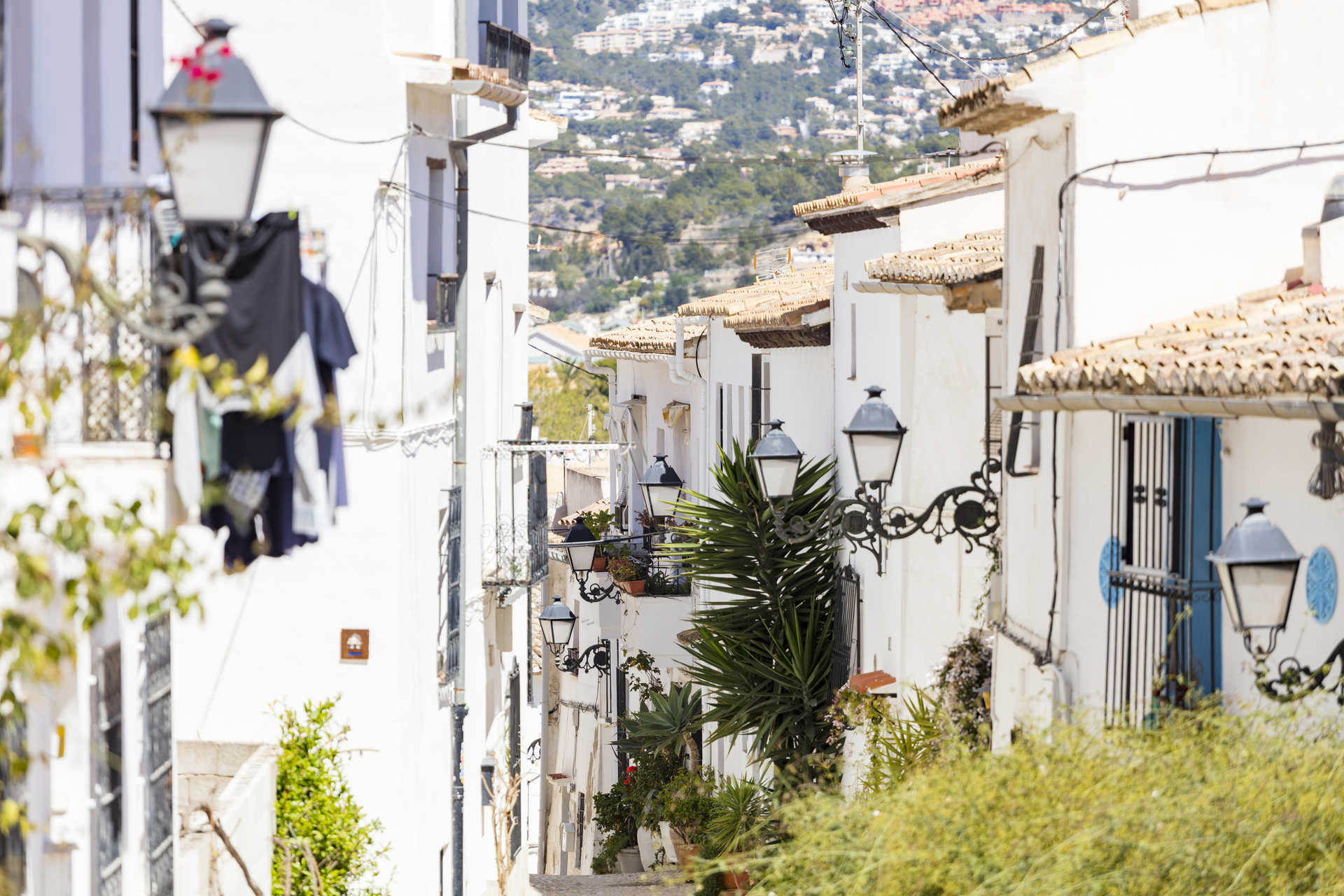 Blick in eine traditionelle schmale Gasse in Altea mit weiß getünchten Fassaden, schmiedeeisernen Laternen und Terrakotta-Ziegeldächern.