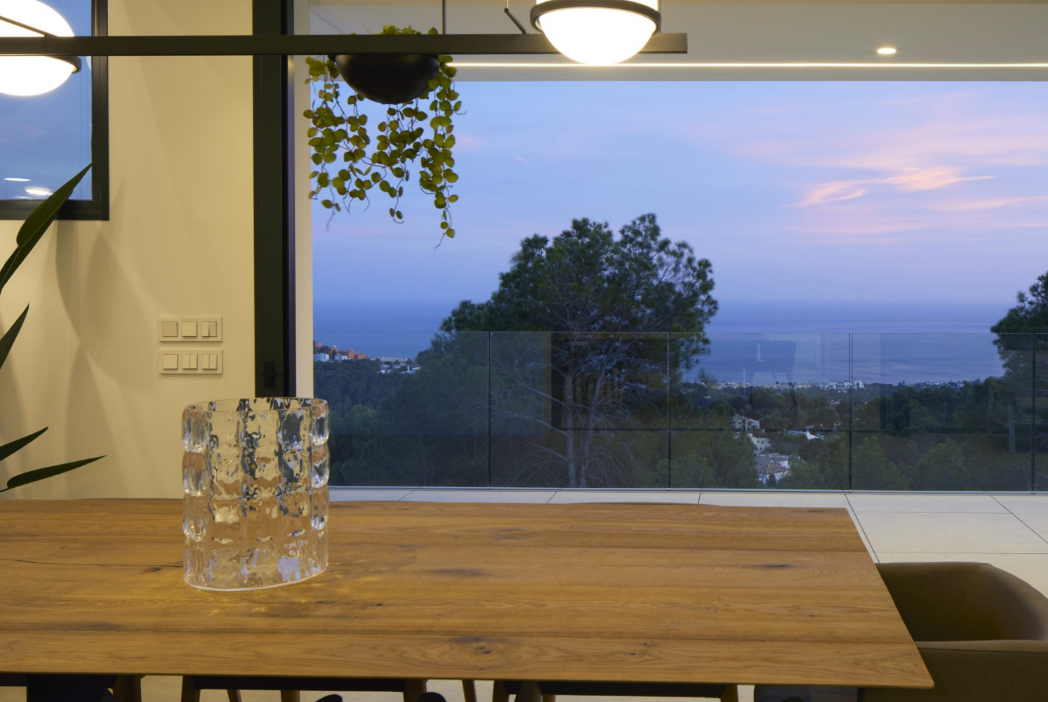 Blick vom Essbereich mit Massivholztisch auf die Terrasse. Bodentiefe Fenster bieten eine Panorama-Aussicht auf das Mittelmeer und die Küste von Altea bei Dämmerung.