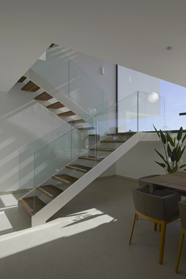 Design-Treppe mit dunklen Holzstufen und Glasgeländer in einem hellen Wohnbereich mit großen Fensterfronten und markantem Lichteinfall.