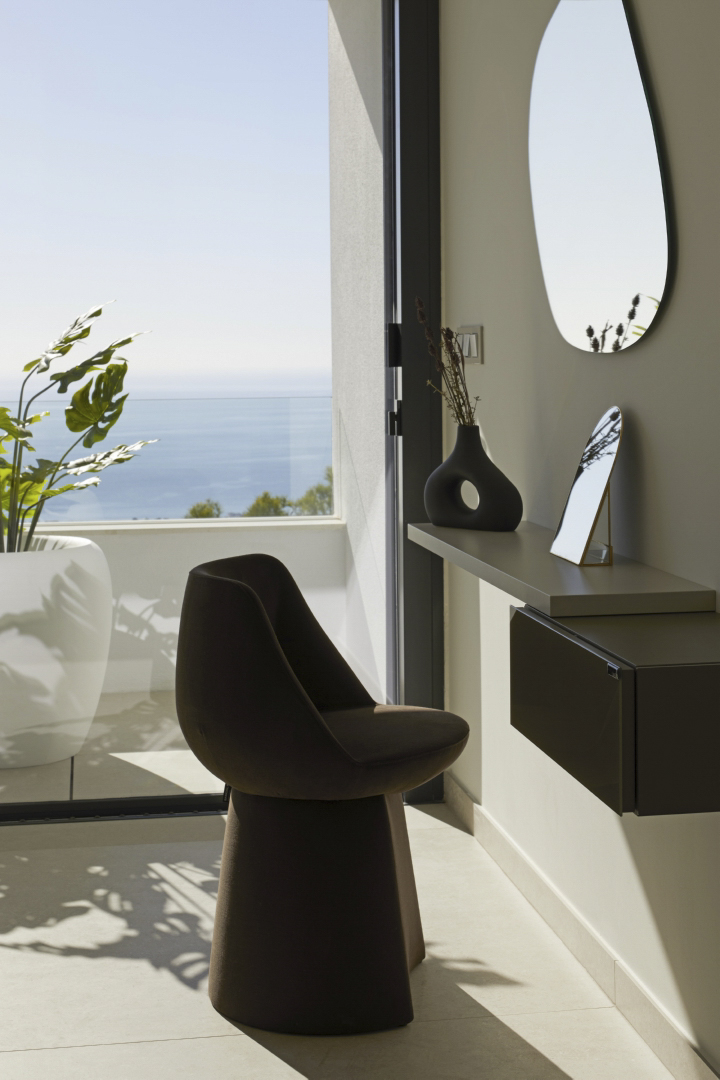 Designer-Schminkbereich mit organisch geformtem Spiegel und skulpturalem Stuhl vor bodentiefen Fenstern mit Blick auf das Mittelmeer.