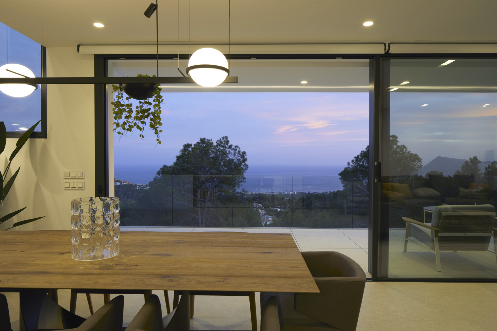 Essbereich mit Massivholztisch und moderner Pendelleuchte, der durch große Glasschiebetüren einen weiten Blick auf das Meer und die Küste bietet.