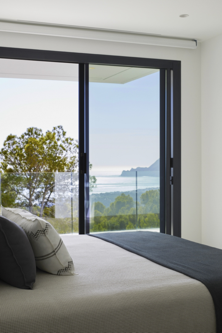 Hauptschlafzimmer mit bodentiefen Glasschiebetüren, dunklen Rahmen und Panoramablick auf das Mittelmeer und die umliegende Berglandschaft.