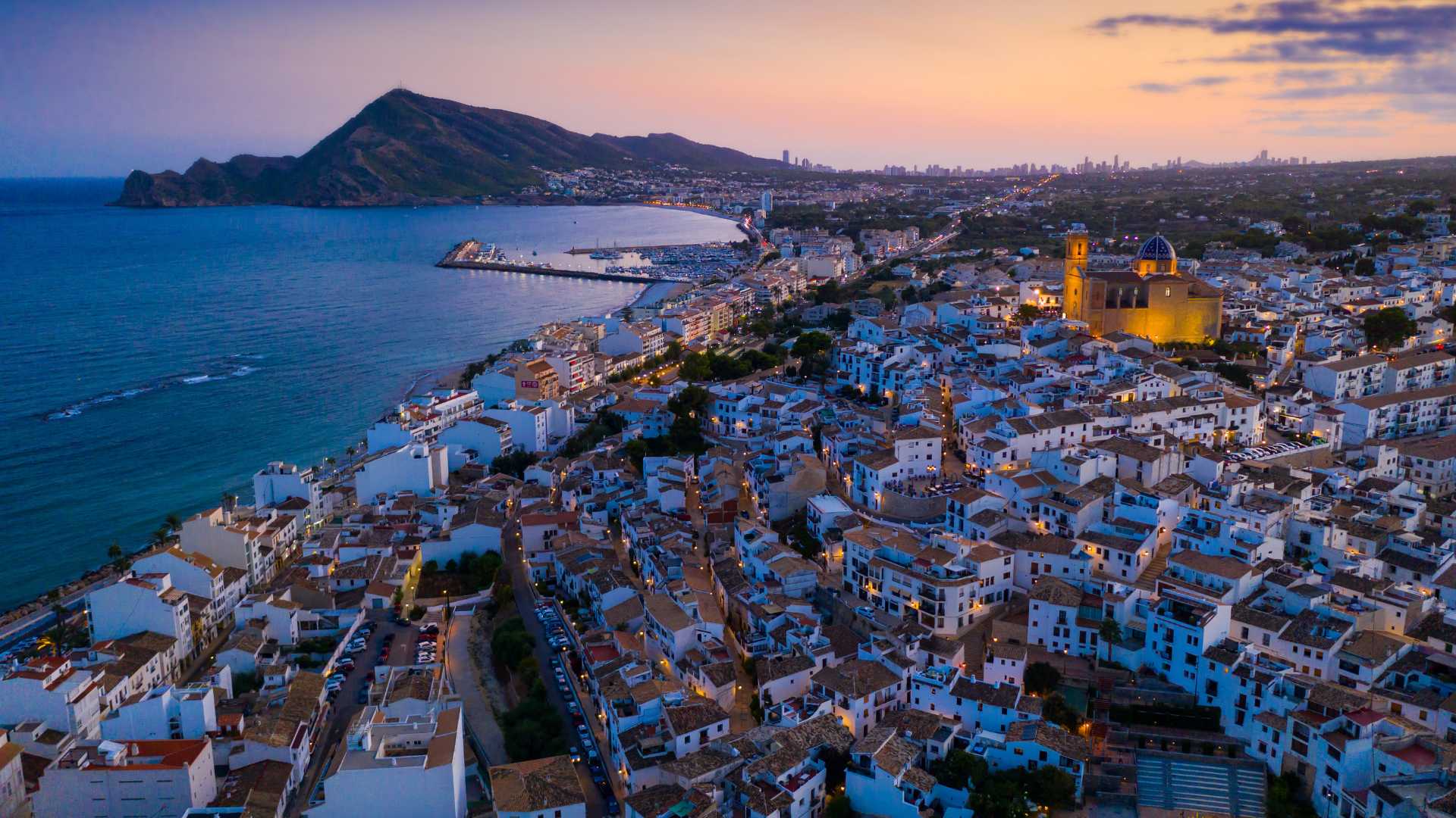 Luftaufnahme von Altea bei Dämmerung mit Blick auf die blau gekuppelte Kirche, die Mittelmeerküste und das Gebirge der Sierra Helada im Hintergrund.