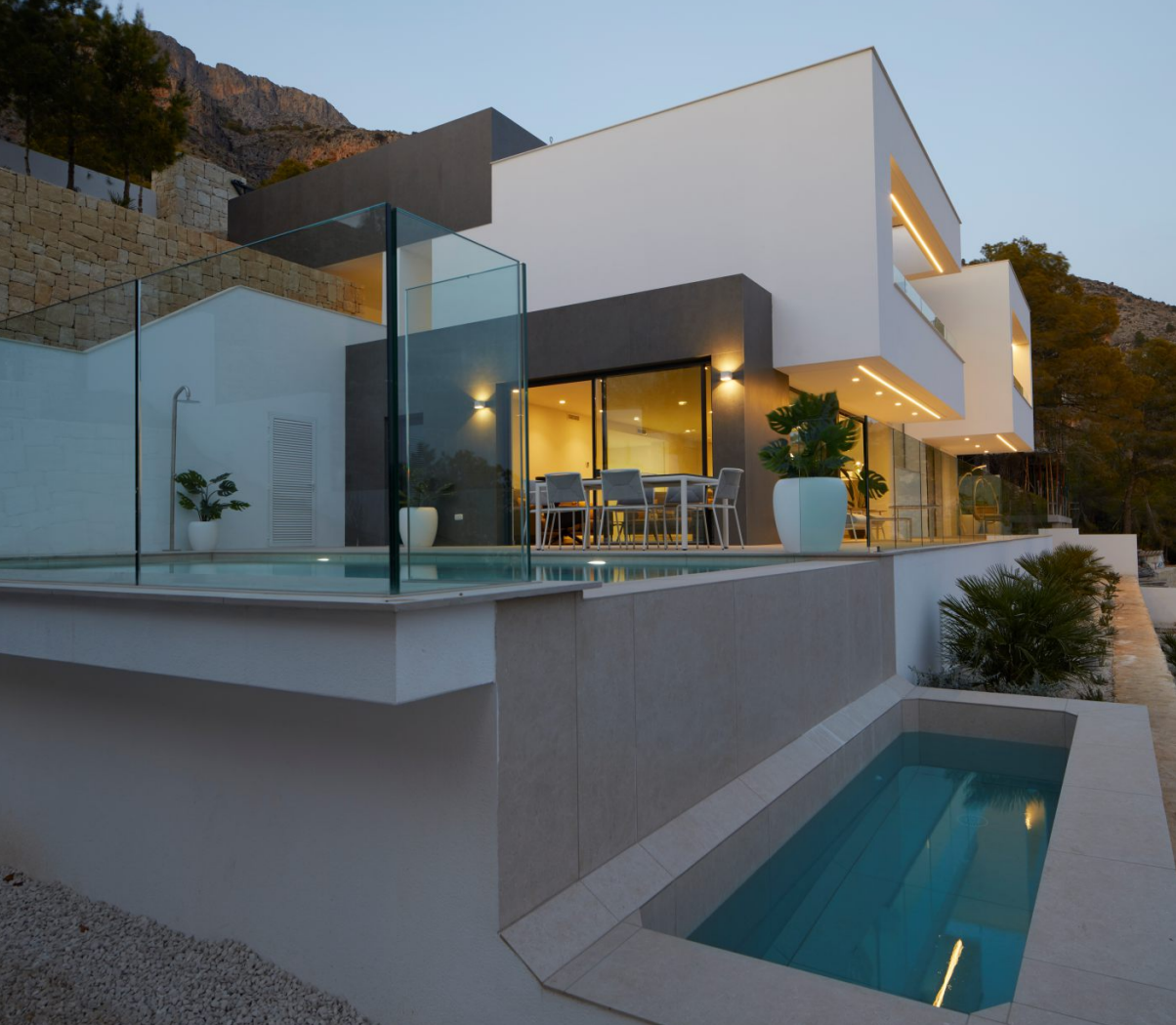 Moderne Außenansicht mit Infinity-Pool, Glasgeländern und beleuchteten Terrassen vor der Kulisse der Sierra de Altea.