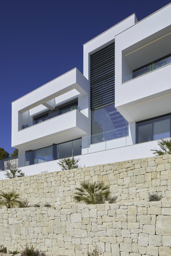 Moderne Fassade mit weißen geometrischen Formen und Glasgeländern über einer Trockensteinmauer mit mediterraner Bepflanzung.