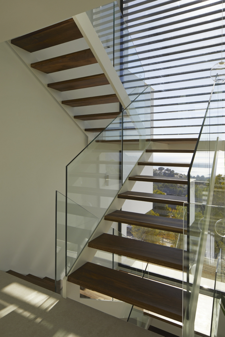 Moderne Treppe mit dunklen Holzstufen und Glasgeländern vor einer großen Fensterfront mit Blick auf die Küste von Altea.