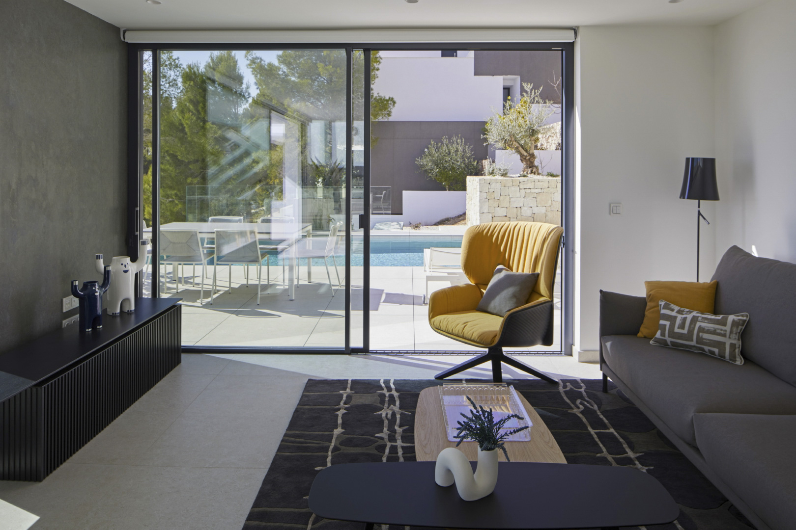 Modernes Wohnzimmer mit großen Glasschiebetüren, die zur Terrasse und zum Infinity-Pool führen. Ausgestattet mit Designermöbeln, einem gelben Sessel und Fliesenboden.