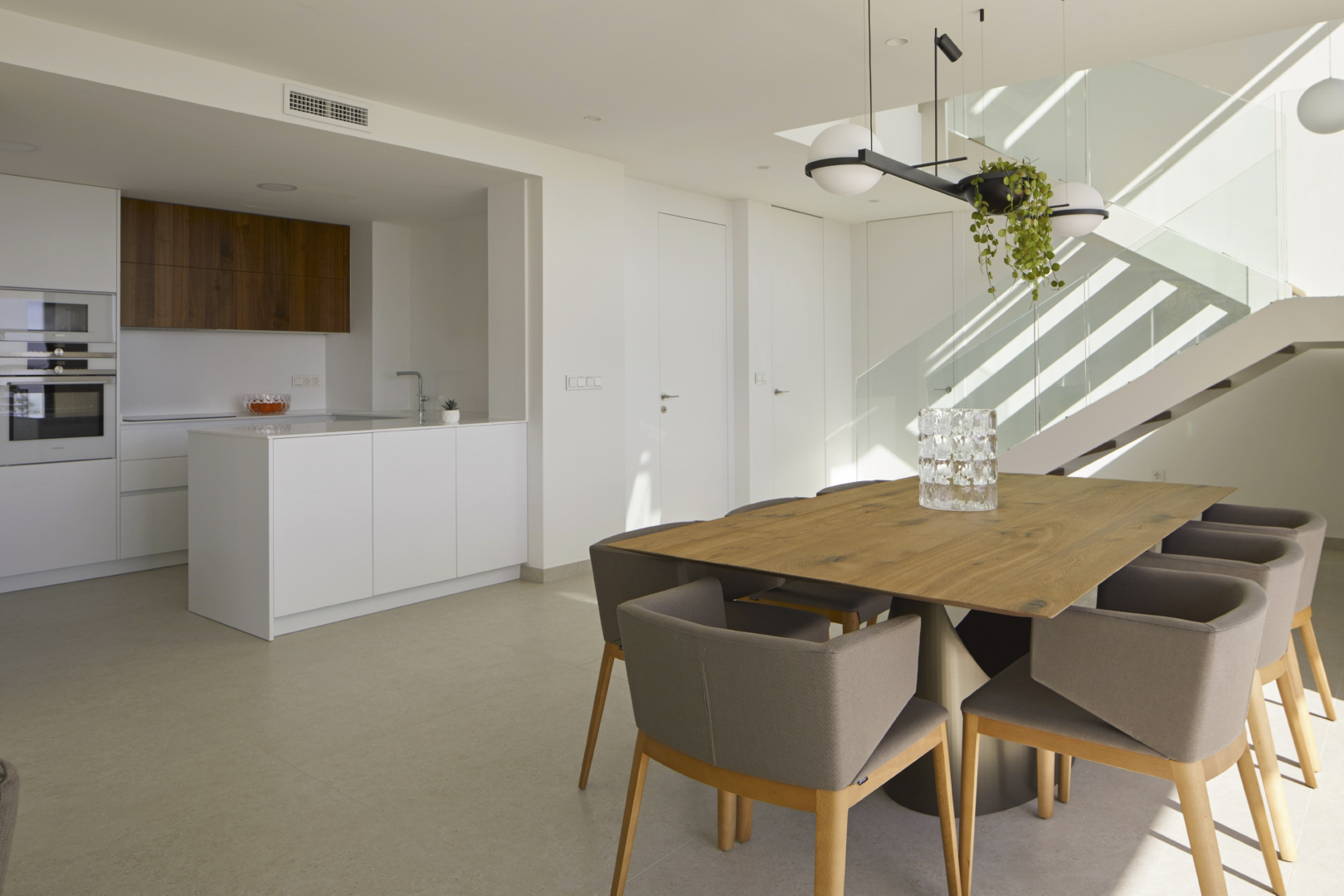 Offener Essbereich mit Massivholztisch, minimalistischer Küche mit weißer Kochinsel und einer modernen Treppe mit Glasgeländer.