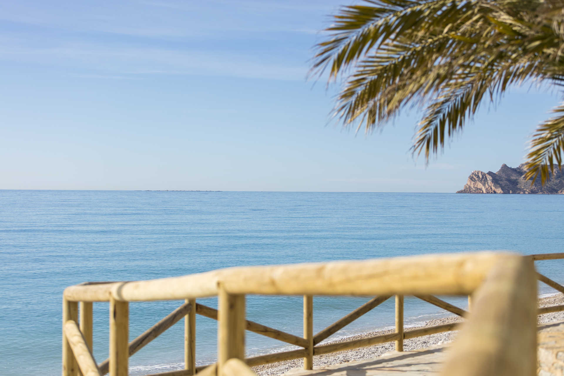Panoramablick auf das Mittelmeer von einem Küstenweg mit Holzgeländer, Palmenzweigen und fernen Felsformationen an der Costa Blanca.