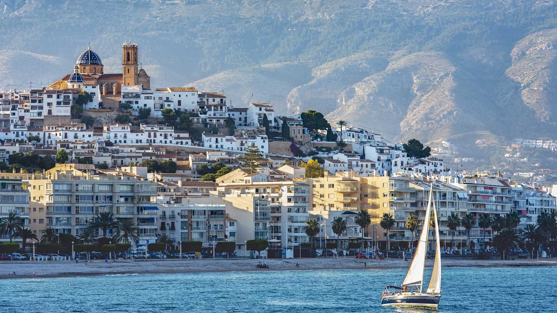 Panoramablick auf die Altstadt von Altea mit der berühmten blau gekachelten Kirche, dem Mittelmeer und einem Segelboot vor der Bergkulisse.